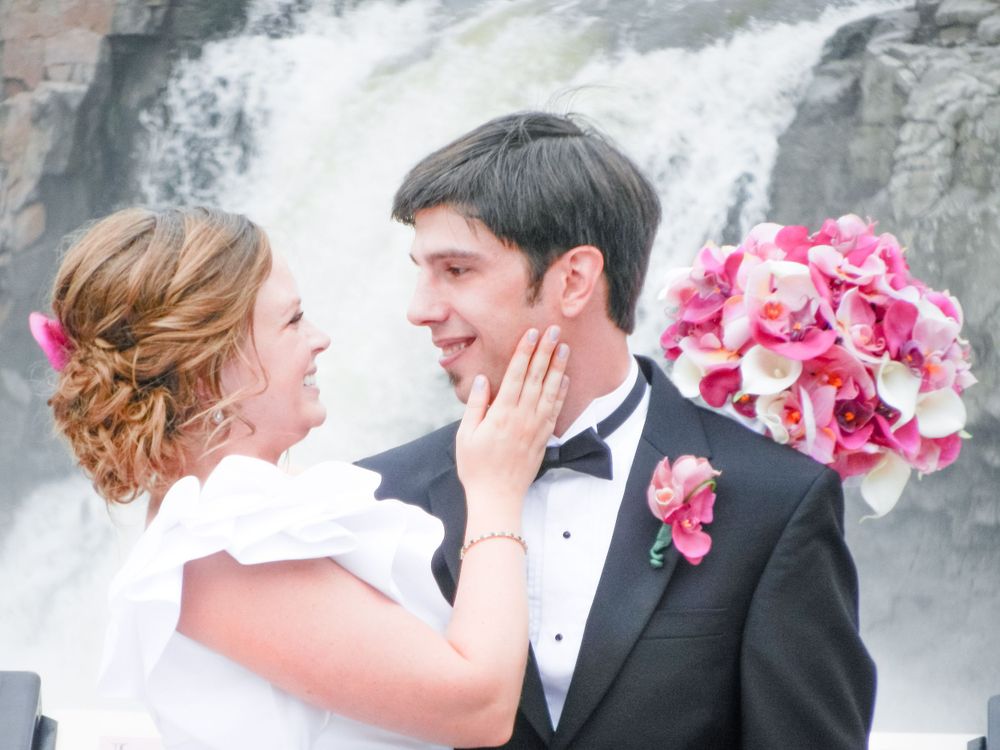 Newlywed couple shares romantic moment by cascading waterfall with pink bouquet.