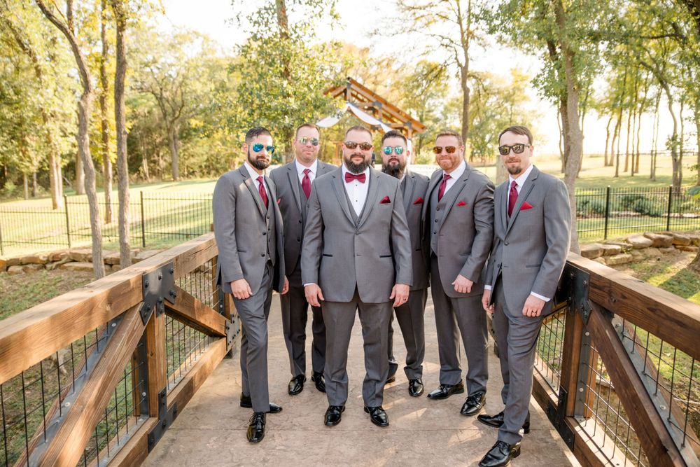 A group wearing matching gray suits and red ties stands together on a wooden bridge surrounded by autumn trees.