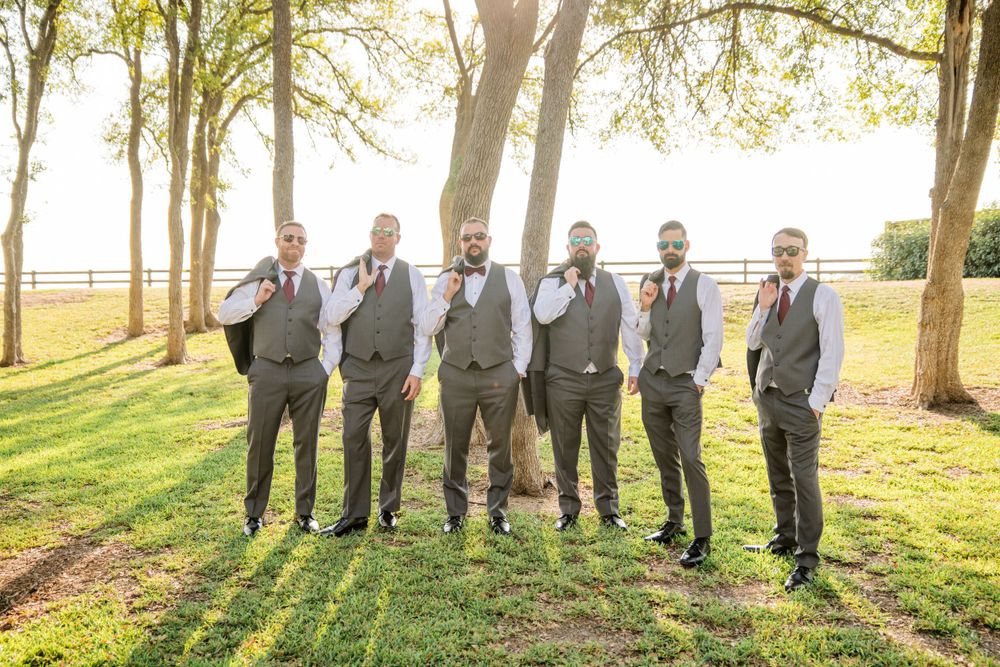 A wedding party in gray vests and black pants poses on a sunlit lawn with trees in the background.