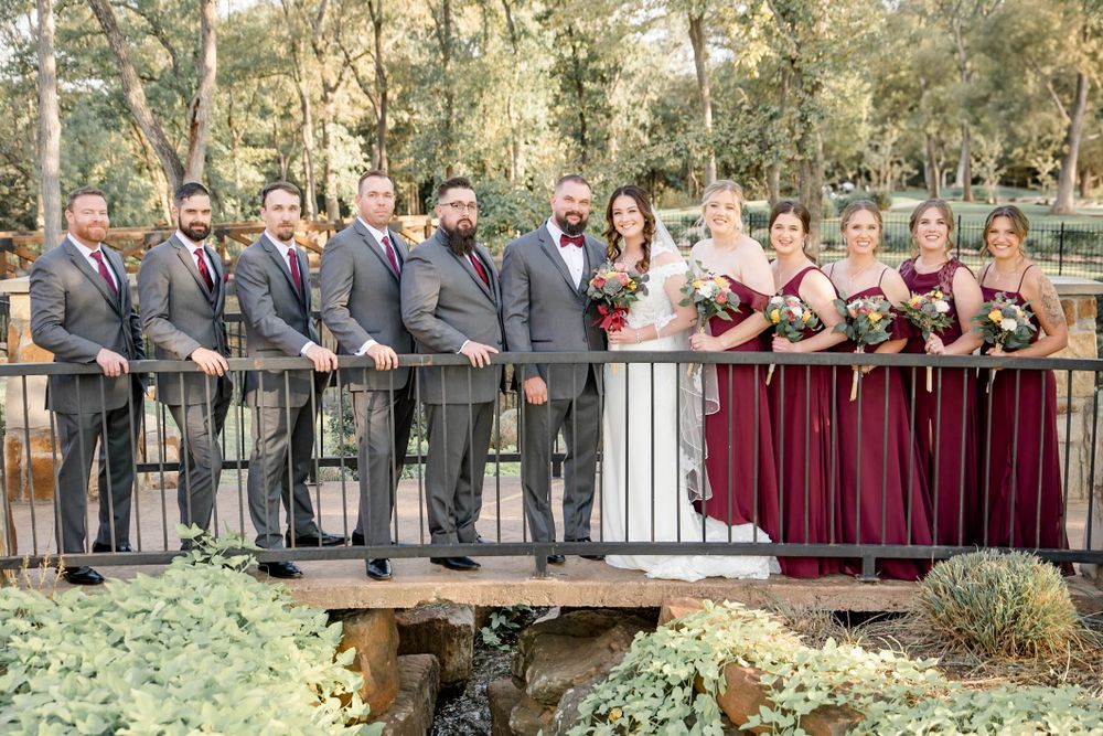 Wedding party in burgundy and gray attire poses together on rustic wooden bridge during outdoor ceremony.