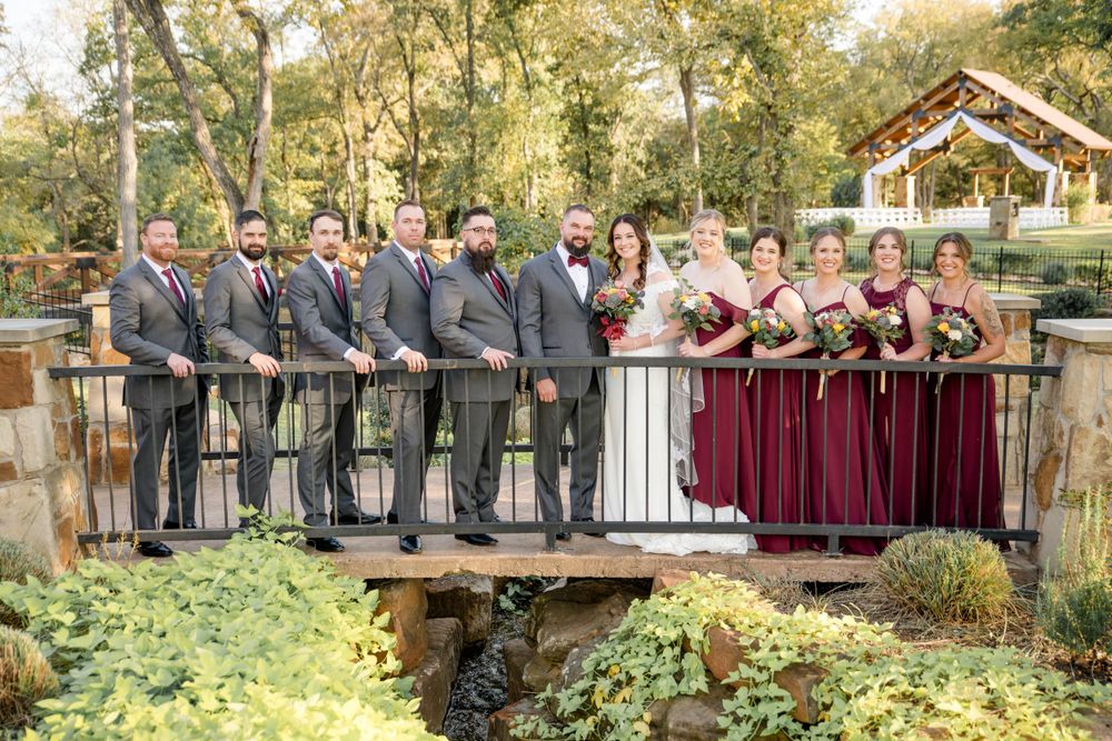 Wedding party in gray suits and burgundy dresses pose for portrait on rustic wooden bridge over stream in garden setting.