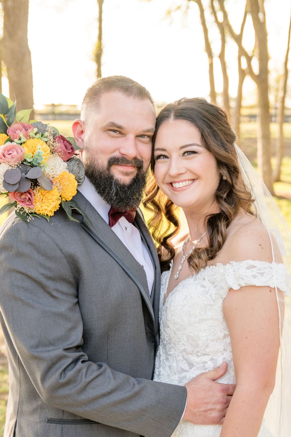 A romantic outdoor wedding photo features the newlyweds embracing with a colorful autumn bouquet during golden hour.