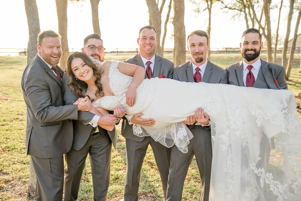 Wedding party wearing gray and burgundy suits playfully lift someone in a flowing white dress outdoors at sunset.