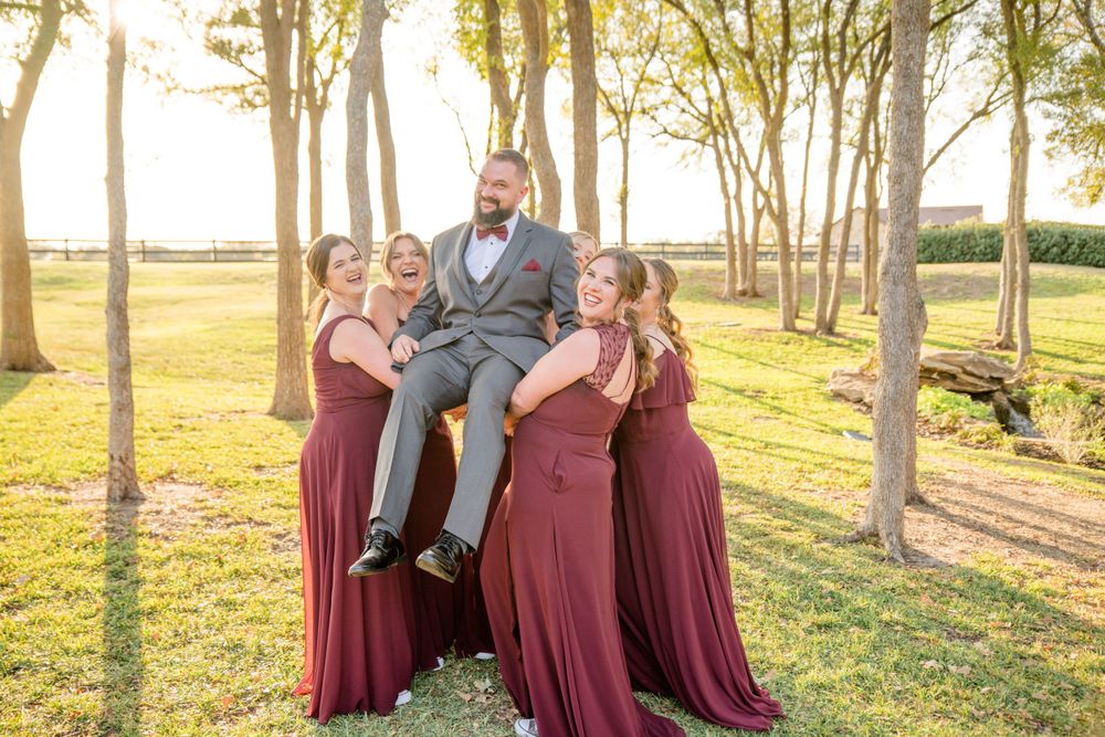 A joyful wedding party poses together in long burgundy gowns among sunlit trees at golden hour.