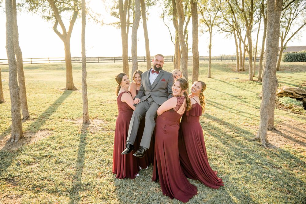 A group in formal attire poses together outdoors among trees during golden hour with burgundy dresses and a grey suit.