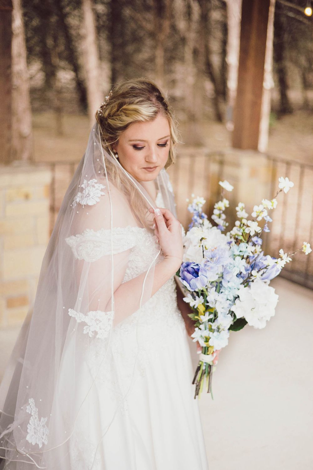A bride in a white lace dress and veil holds a beautiful bouquet of blue and white flowers in a wooded setting.