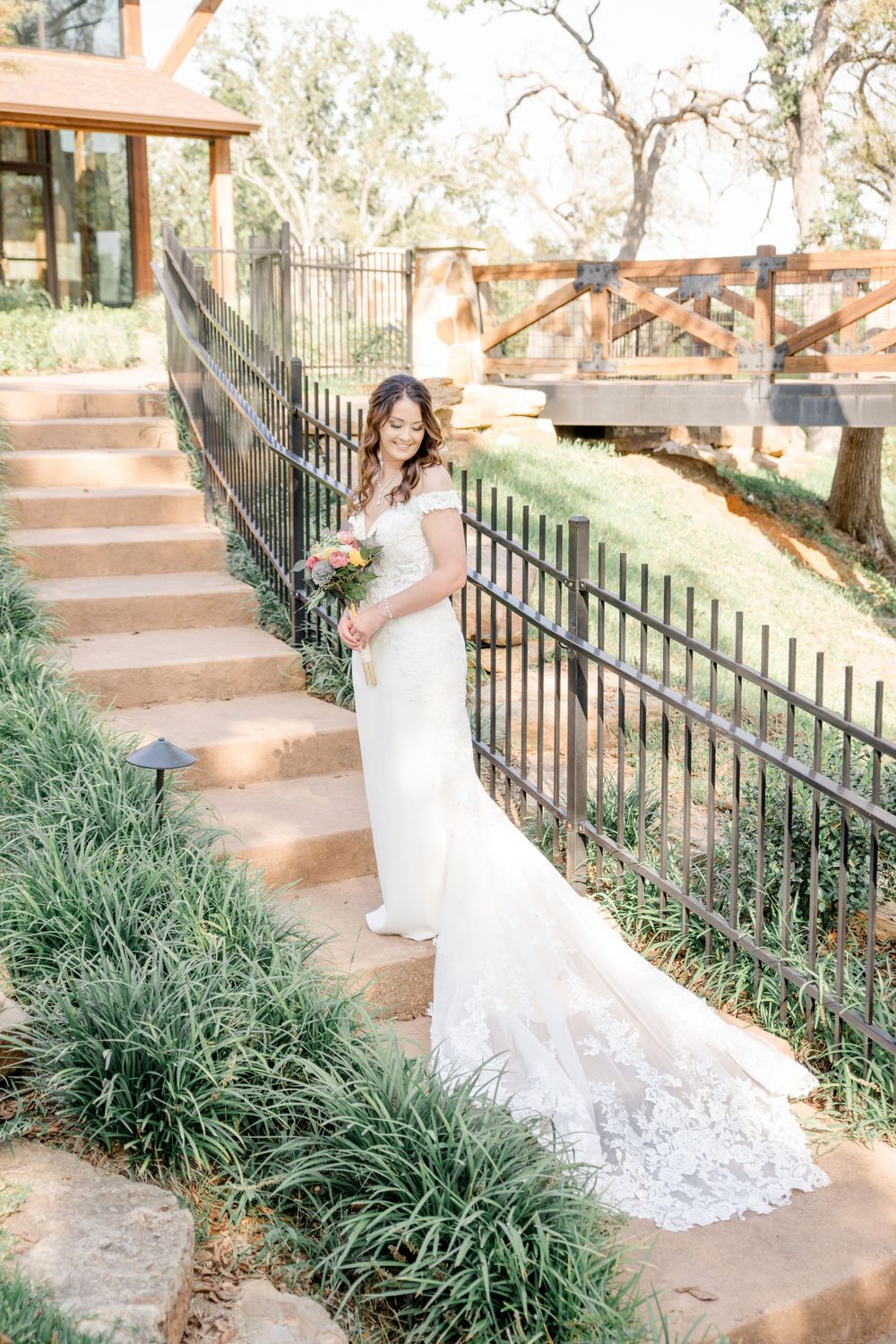 White wedding gown with lace train sweeps across stone steps bordered by greenery and ornate iron railing.