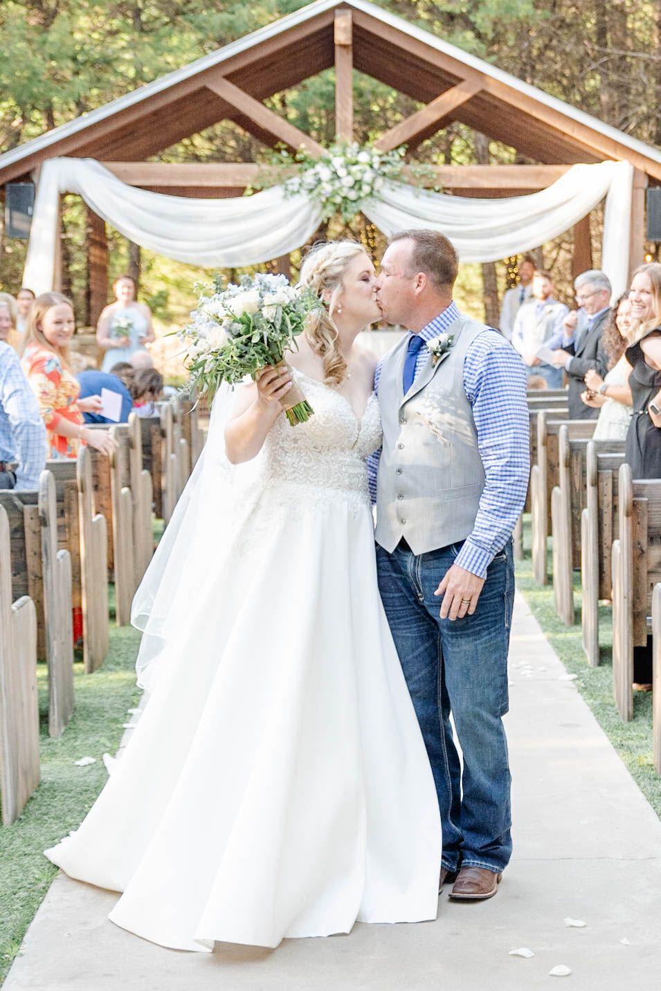 A romantic wedding ceremony under a wooden arch decorated with white drapes and flowers in an outdoor forest setting.