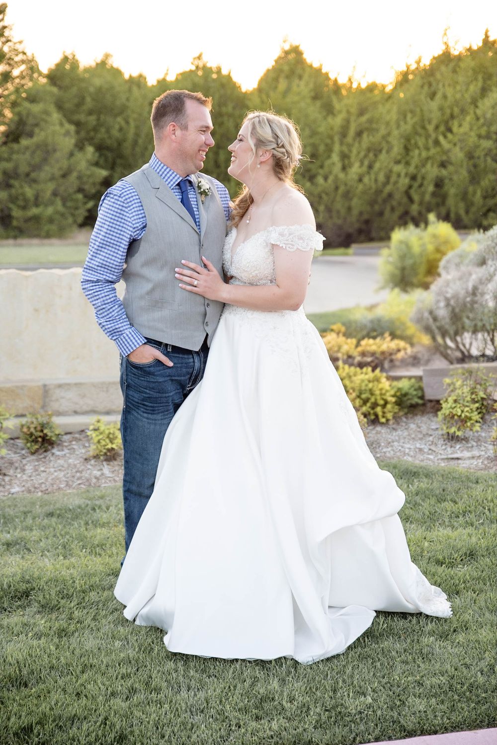 A romantic outdoor wedding scene at sunset with a couple in casual-formal attire embracing on a manicured lawn.