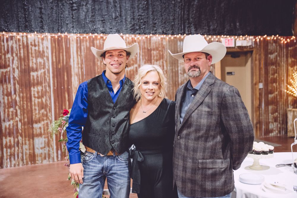 Three people stand together smiling at an outdoor event venue with rustic wooden fence and string lights in background.