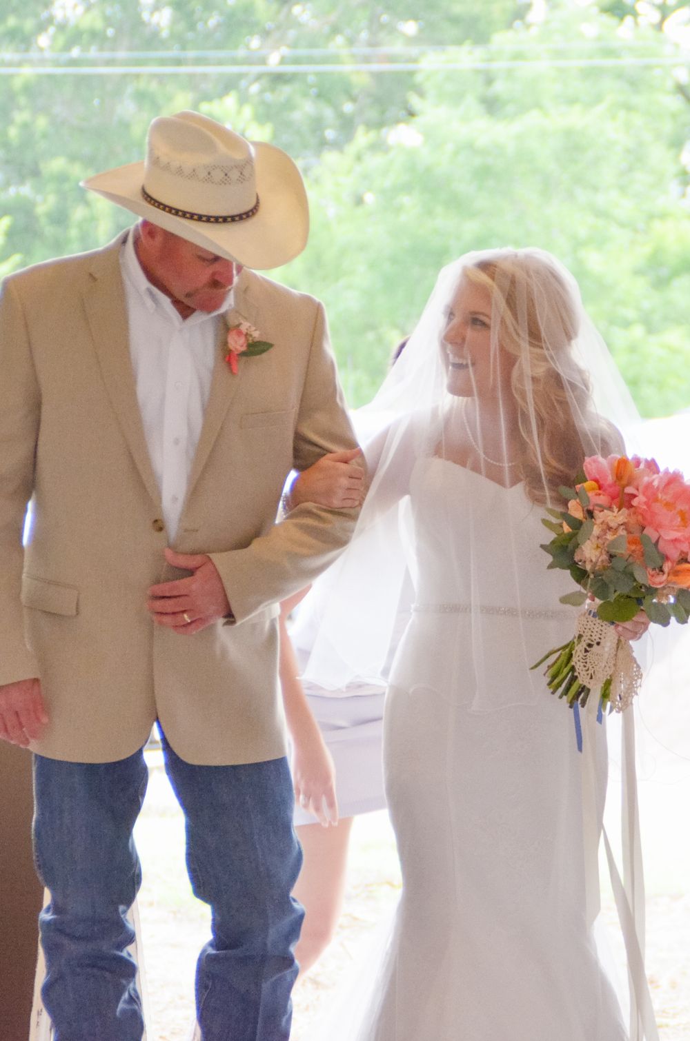 A bride in a white dress walks with a companion wearing a cowboy hat and tan blazer during a wedding ceremony.