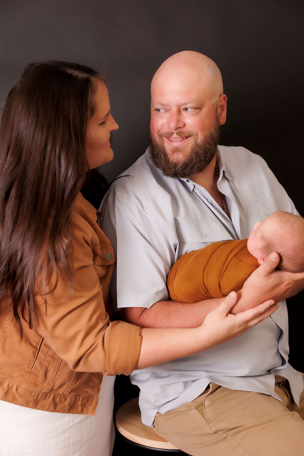 A young couple in casual attire gazes lovingly at their newborn baby swaddled in brown fabric.