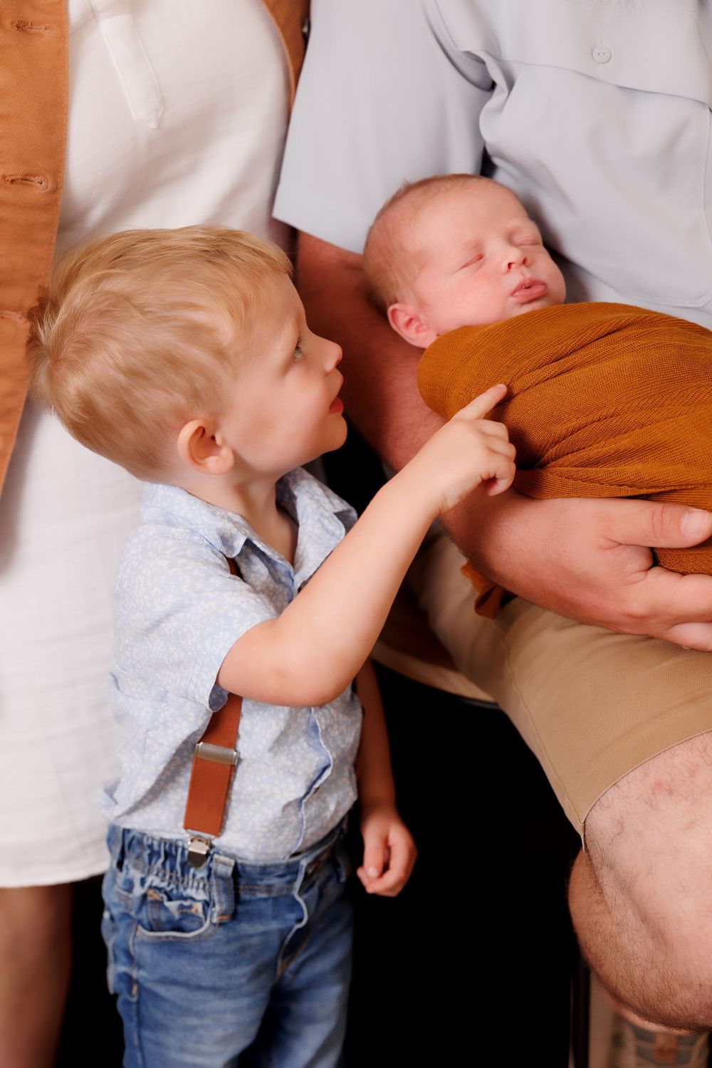 A toddler in denim points at a sleeping newborn baby wrapped in brown fabric.
