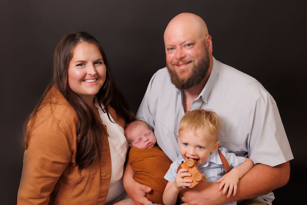 A family portrait featuring parents with their toddler and newborn against a dark background.