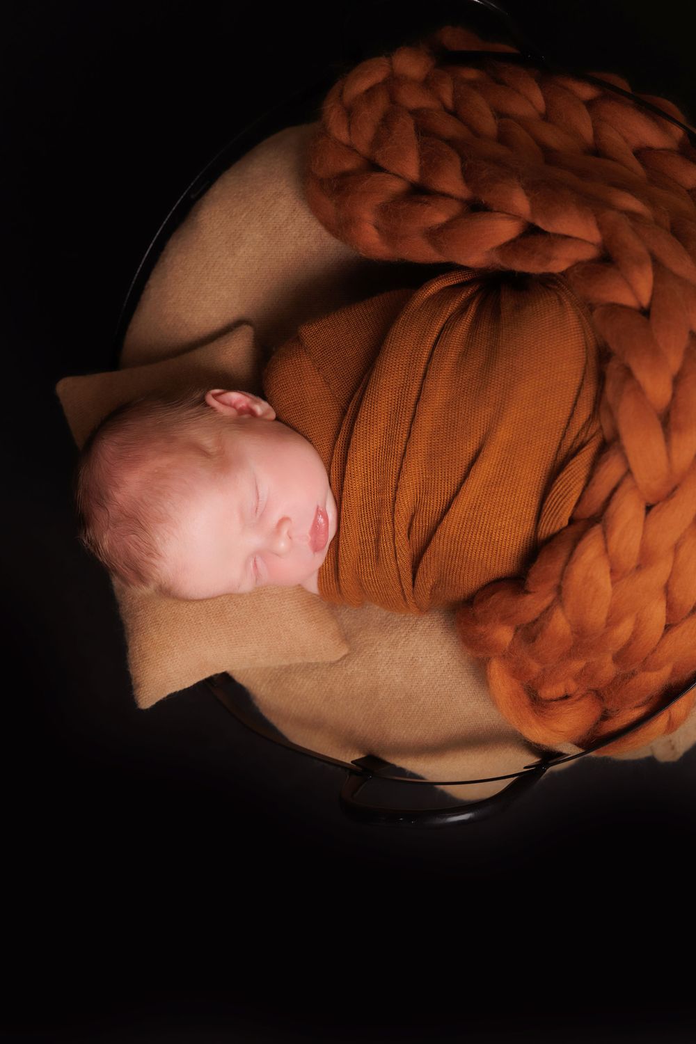 Newborn sleeping peacefully wrapped in soft brown knitted blanket against dark background.