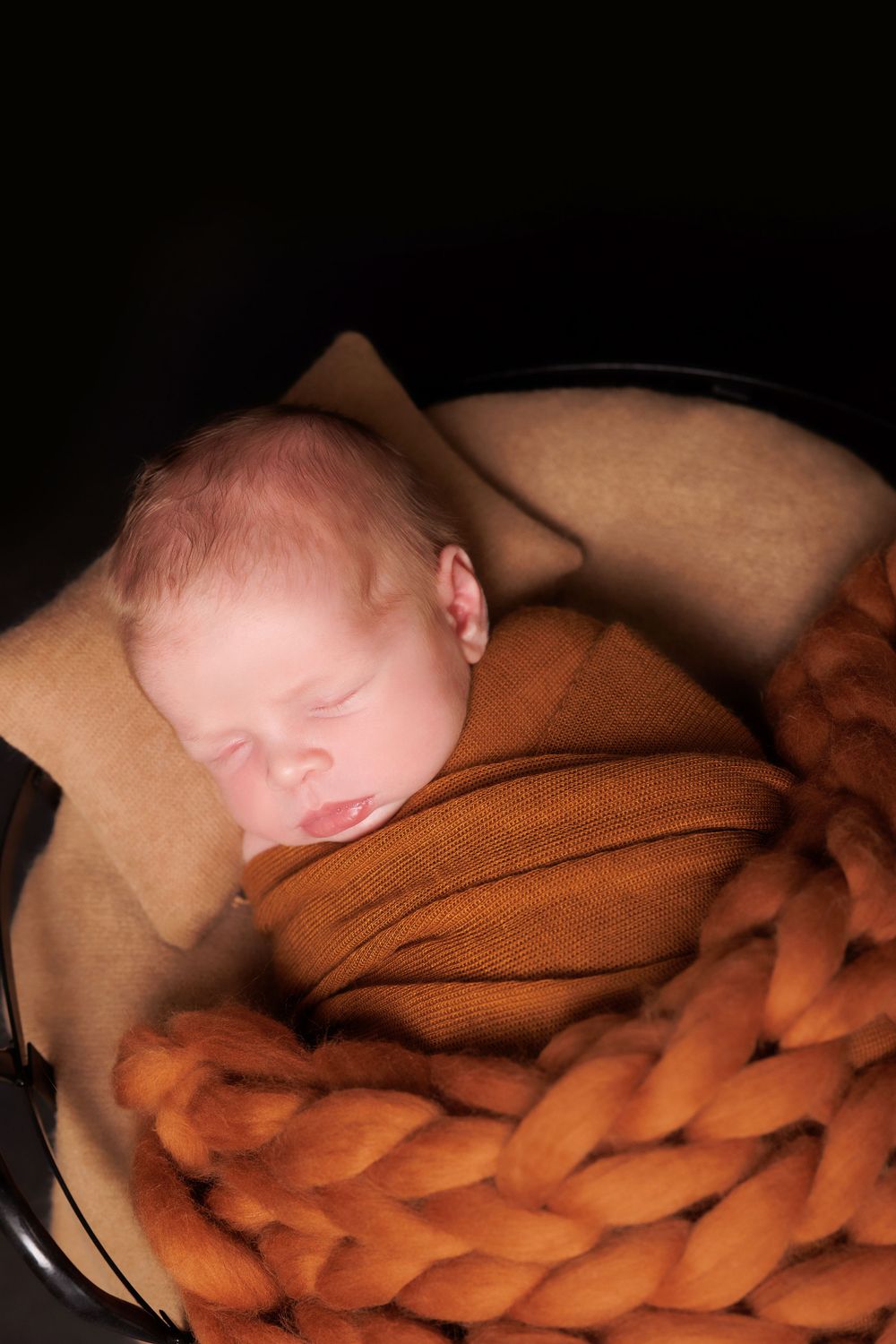 Peaceful sleeping newborn rests on soft brown knitted blanket and tan pillow with warm lighting.