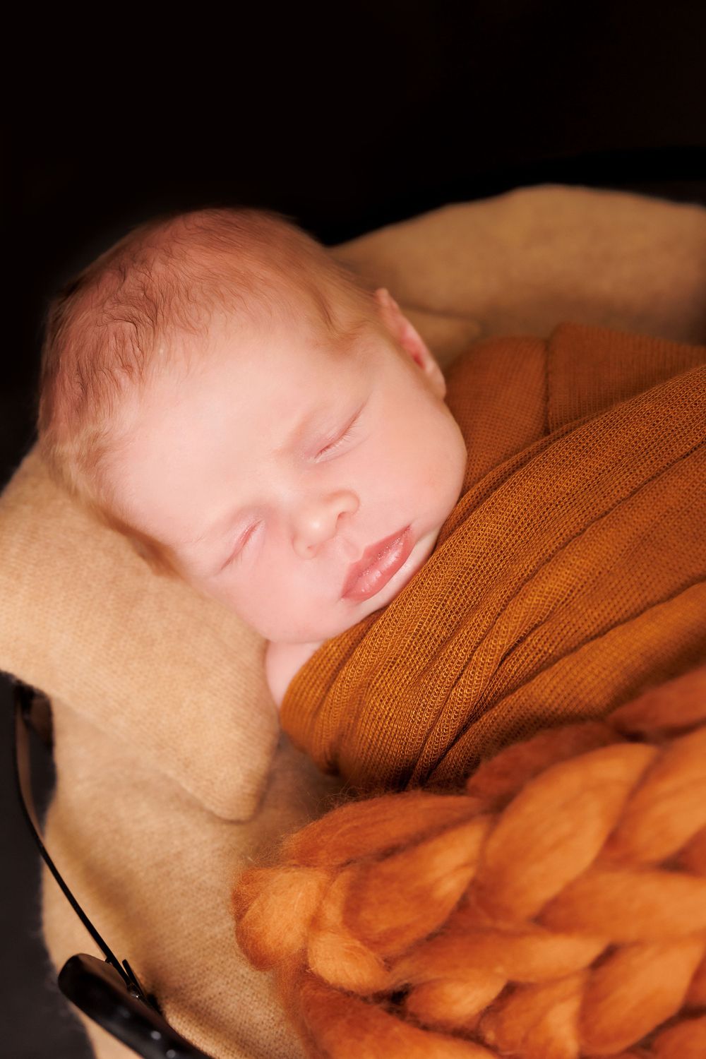 A sleeping baby nestled in orange and beige blankets in dim lighting.