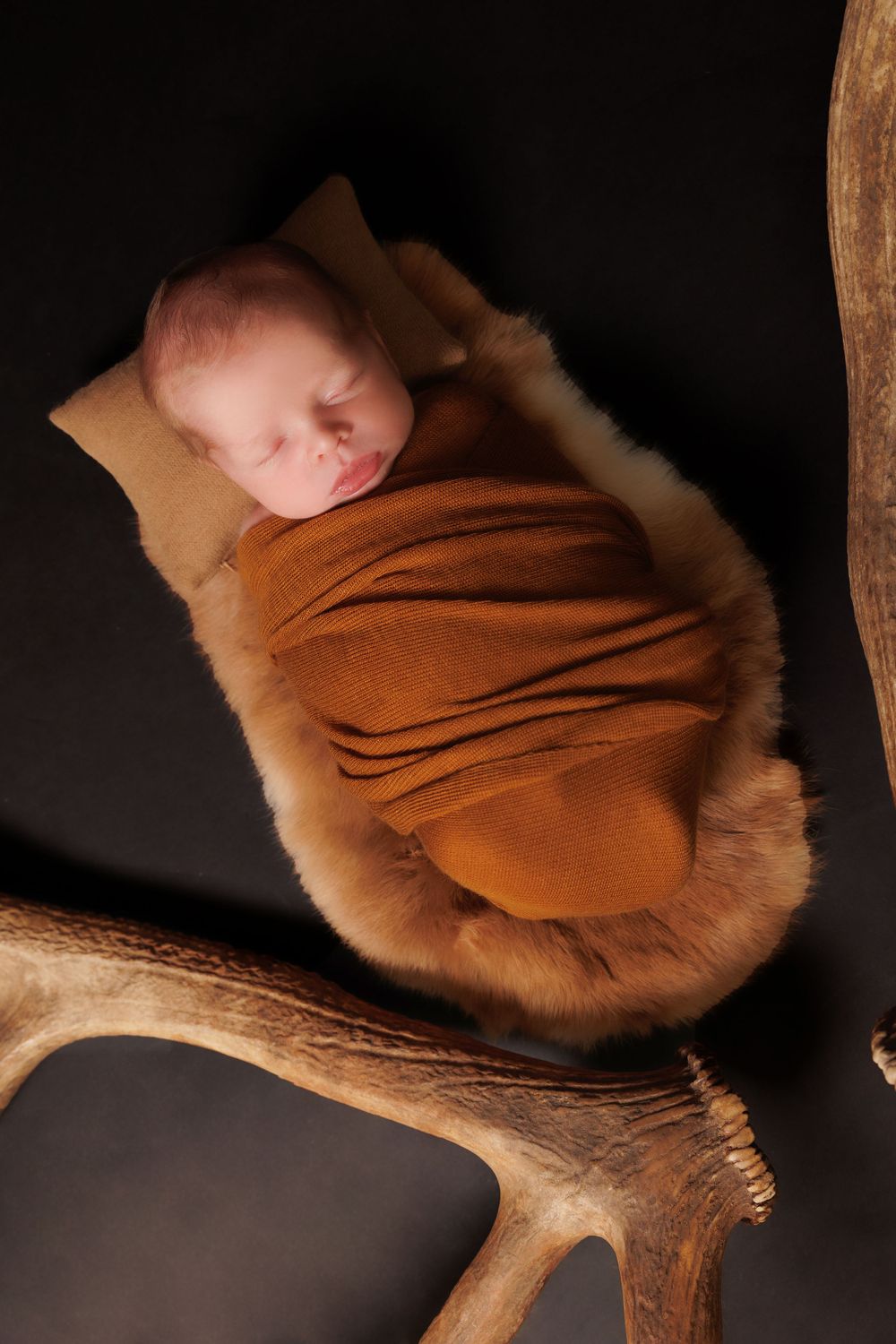 A sleeping newborn wrapped in brown fabric lies on a furry surface next to an antler.