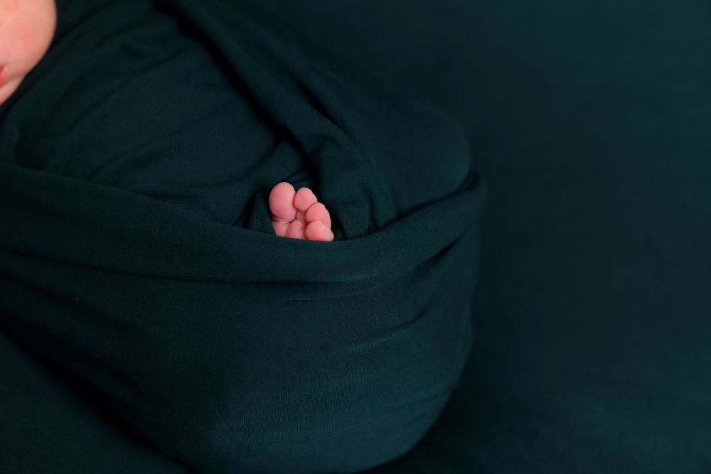 A tiny newborn baby foot peeks out from dark green swaddling blanket.