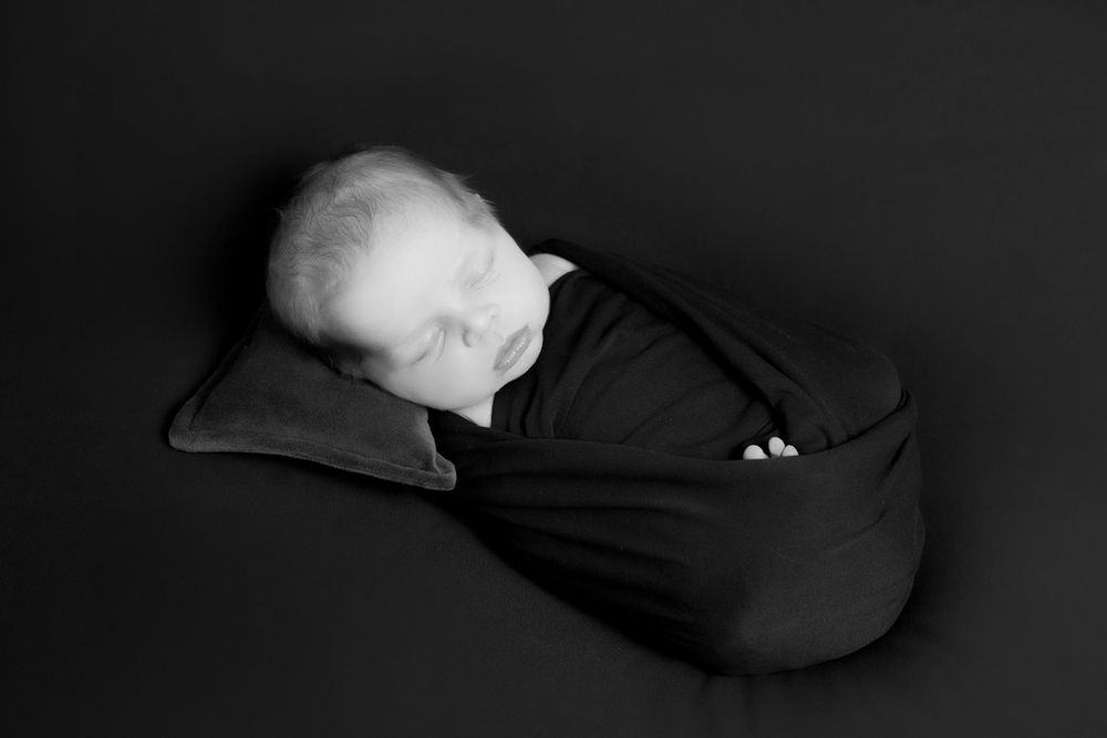 A sleeping newborn baby wrapped in a black swaddle against a dark background.