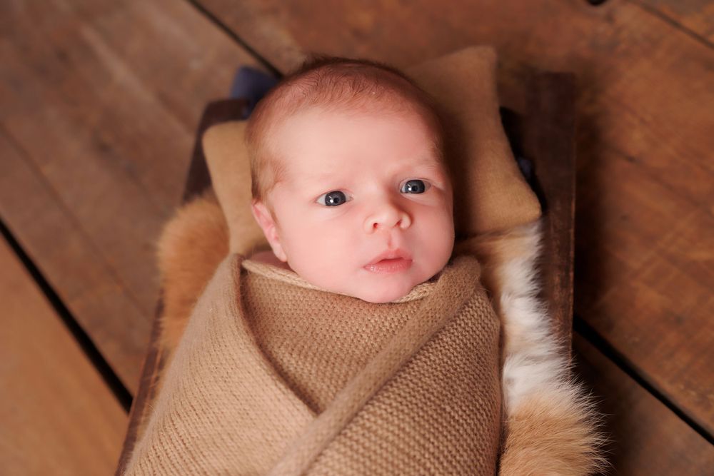 Baby wrapped in soft brown blanket lying on wooden floor with gentle expression.