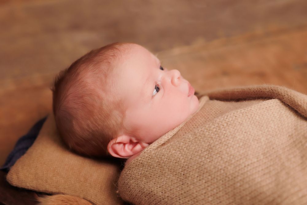 Newborn baby wrapped in tan knit blanket gazes upward against brown backdrop.
