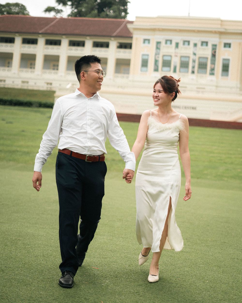 Couple in wedding attire strolling hand in hand on manicured lawn with historic building backdrop.