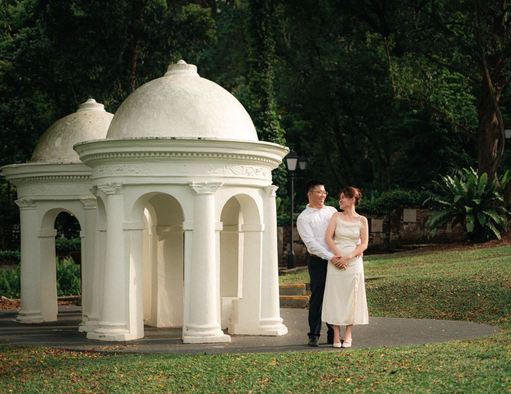 Wedding couple posing beside white domed gazebos in a lush garden setting.