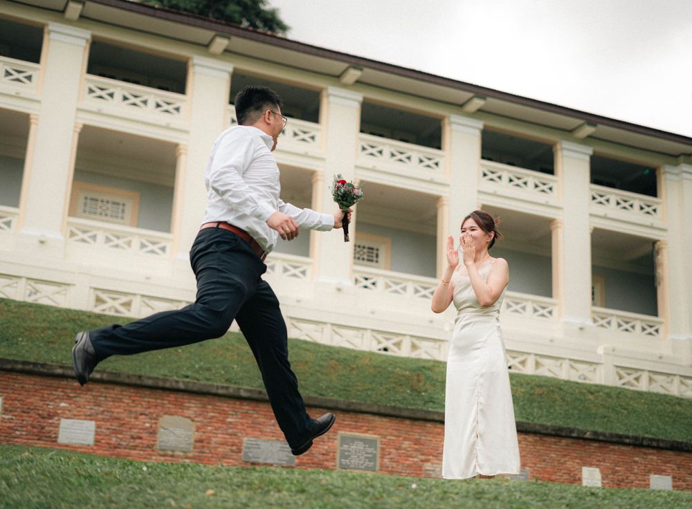 Energetic wedding photo with groom jumping on hill while bride watches below.