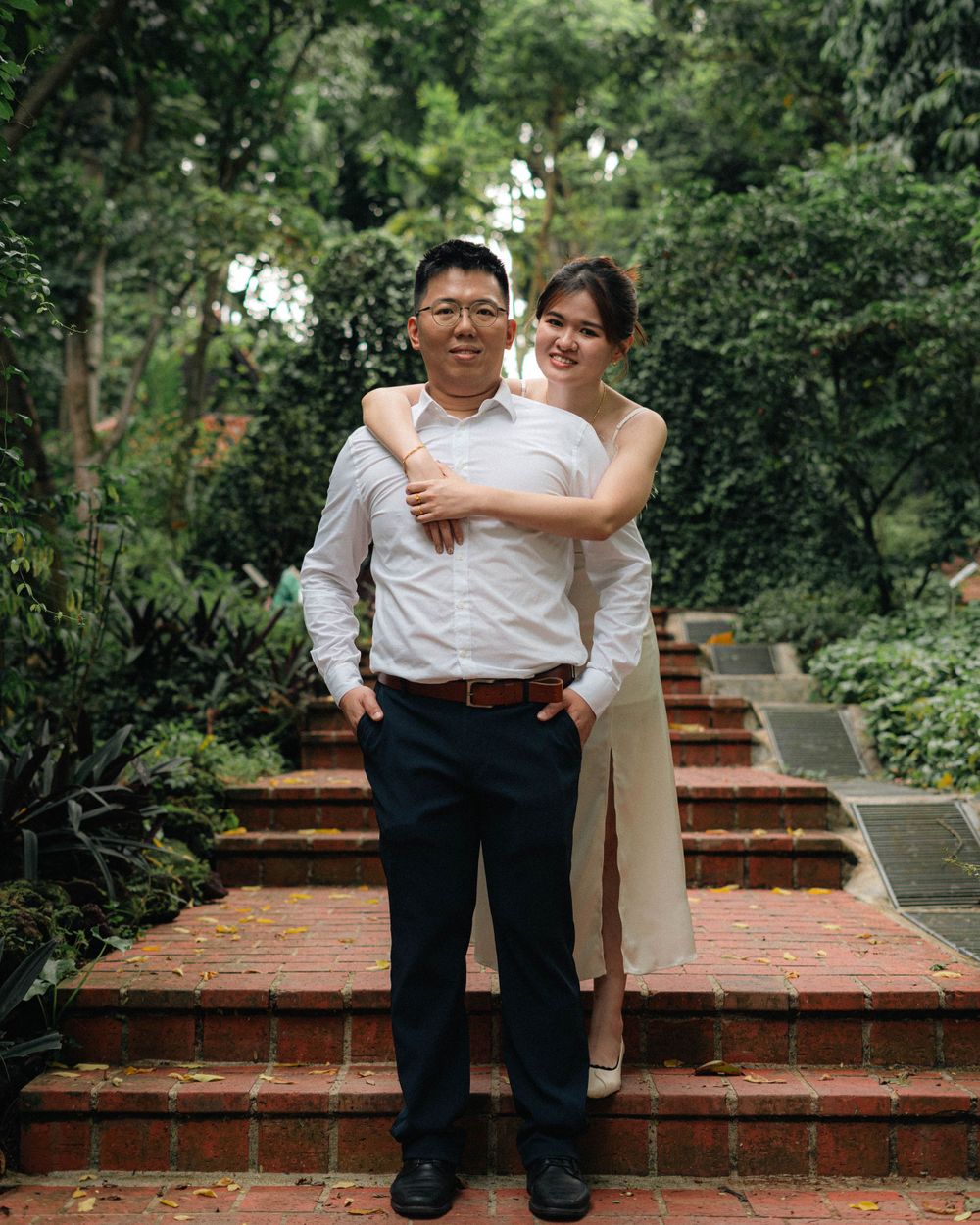 A couple embraces on brick steps surrounded by lush greenery in an outdoor garden setting.