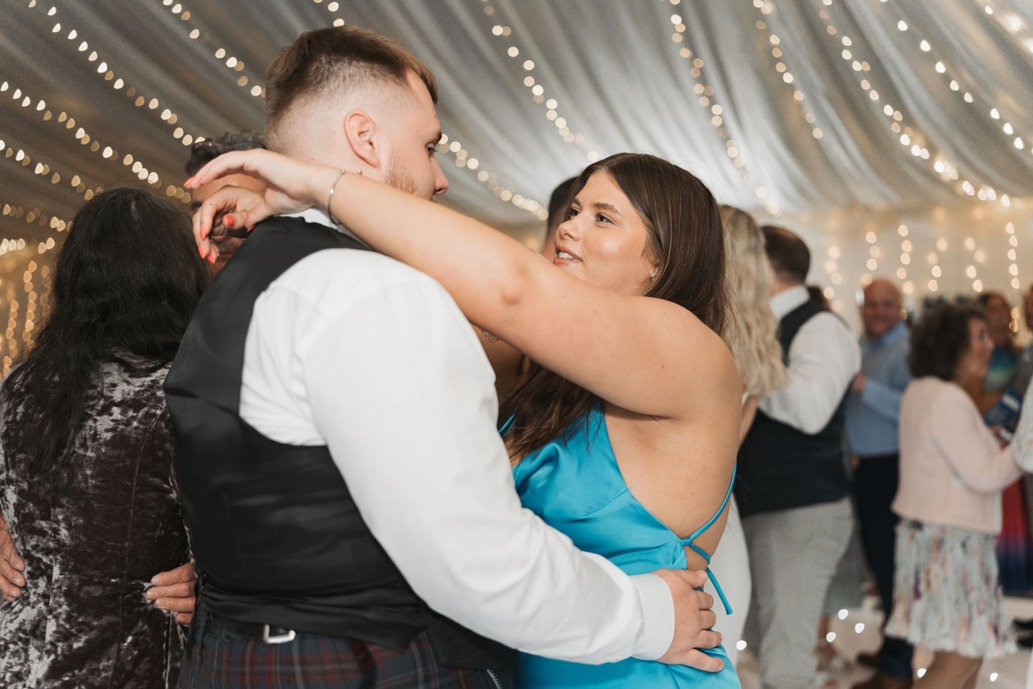A couple dancing together at a wedding reception under twinkling fairy lights with guests in the background.