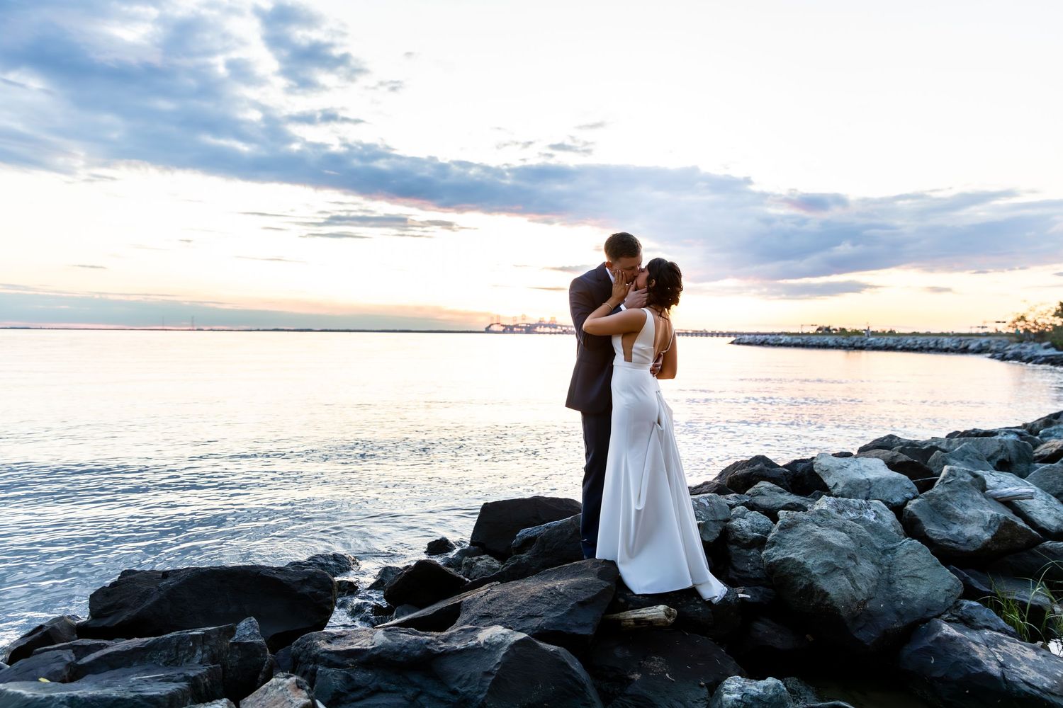 A couple shares a romantic moment on rocky coastal shores of Maryland at sunset with water and cloudy skies in the background.