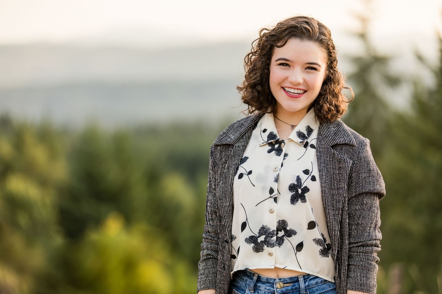 A person in a gray cardigan and floral top stands outdoors smiling with a scenic mountain view in the background.