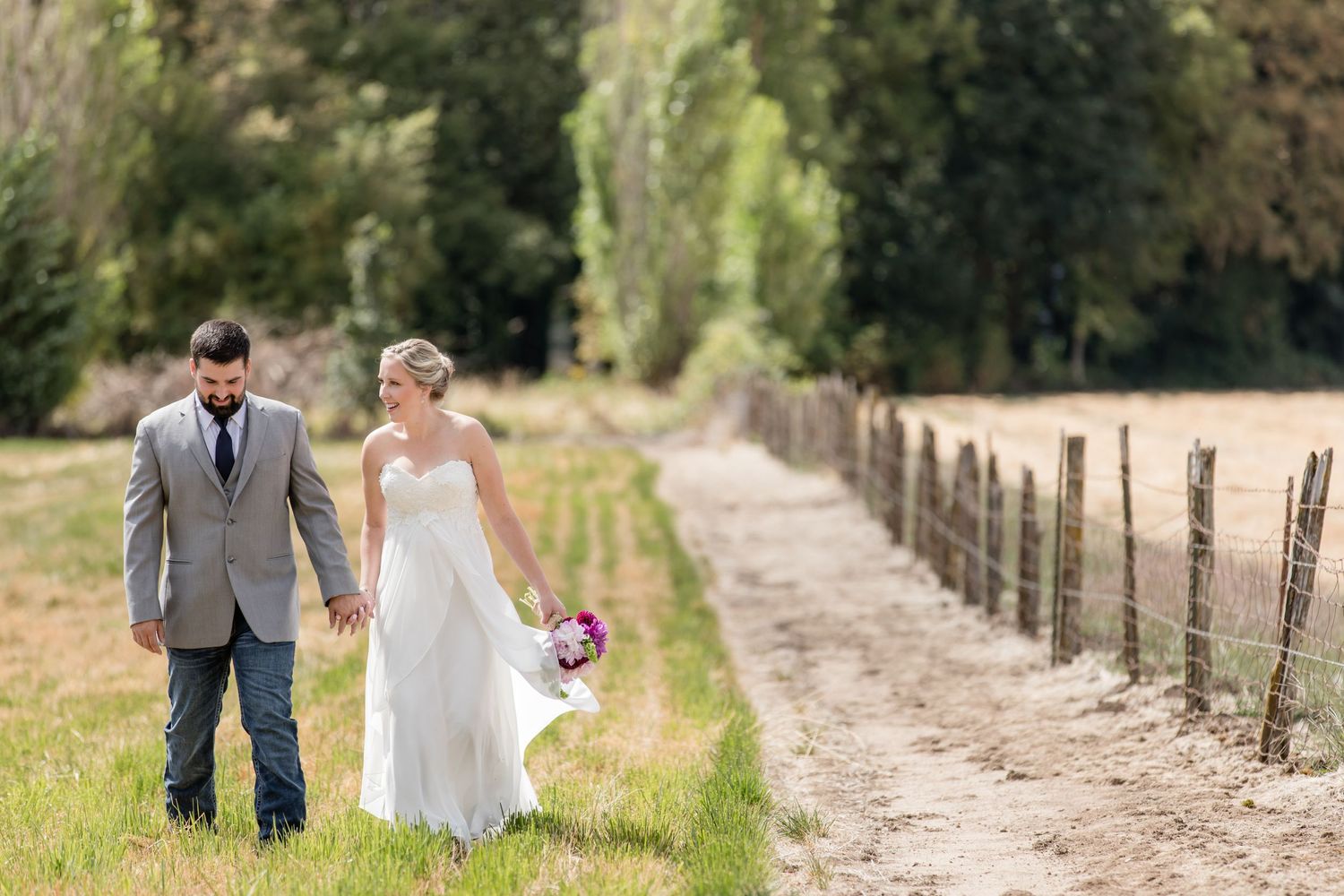 Wedding couple walking on a rustic dirt path lined with wooden fencing in a countryside setting.