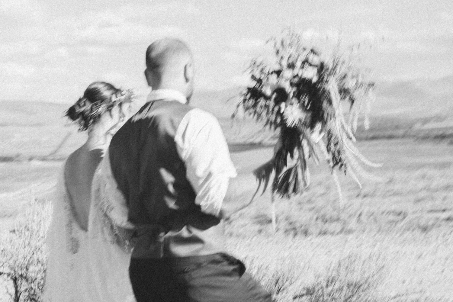 Black and white wedding photo showing couple in field holding rustic flower bouquet.