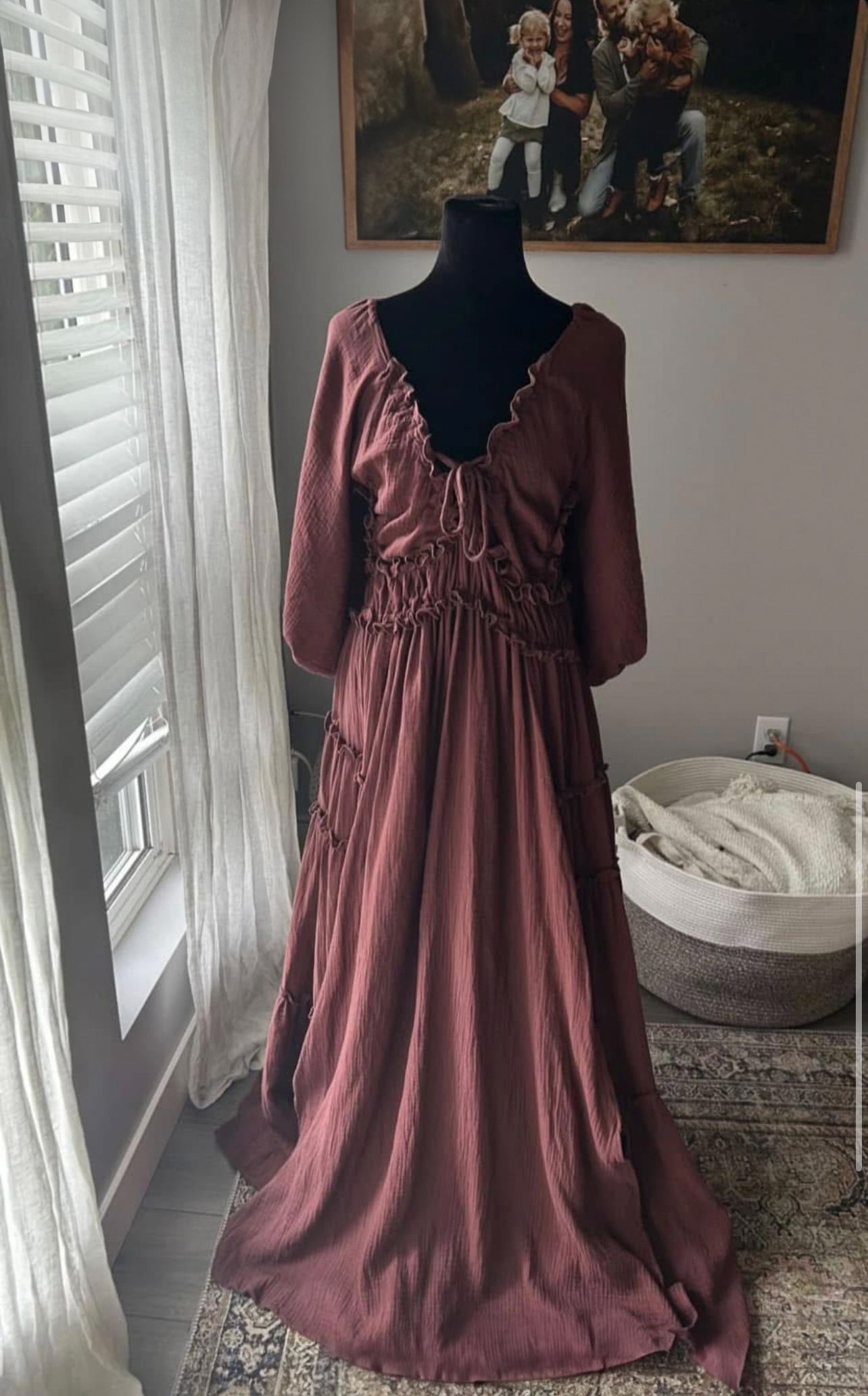 A dusty rose colored romantic maxi dress with ruffled bodice and long sleeves displayed on a black mannequin near window.