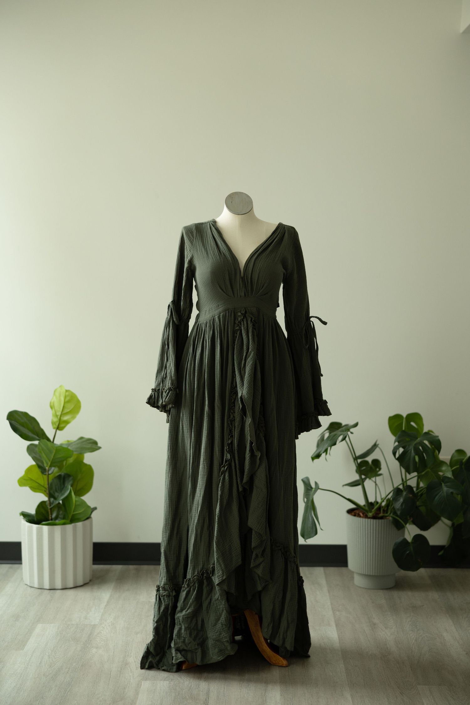 Floor-length black romantic dress with bell sleeves and tiered skirt displayed on mannequin beside houseplants.