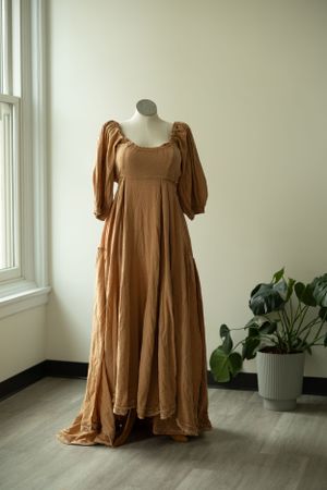 A flowing caramel-colored empire waist maxi dress with puff sleeves displayed on a mannequin near a window and potted plant.