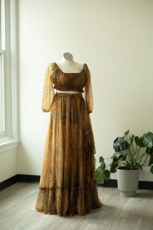 A vintage brown floral maxi dress with sheer puffy sleeves on a mannequin next to a potted monstera plant.