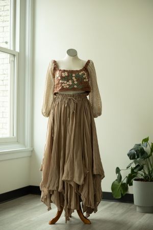 Bohemian brown dress with floral embroidery and flowing asymmetrical skirt displayed on mannequin near window and plant.