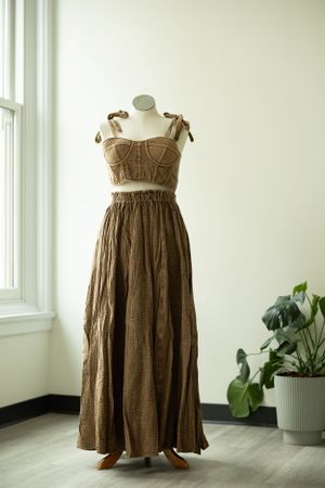A brown two-piece summer outfit with a cropped bustier top and flowing maxi skirt displayed on a mannequin near a window.