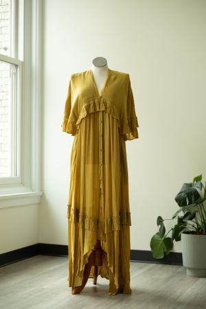 A flowy mustard yellow maxi dress with tiered ruffles and elbow-length sleeves displayed on a mannequin near a window.