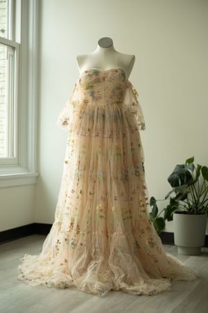 A romantic sheer tulle gown with floral embroidery displayed on a mannequin near a window and potted plant.