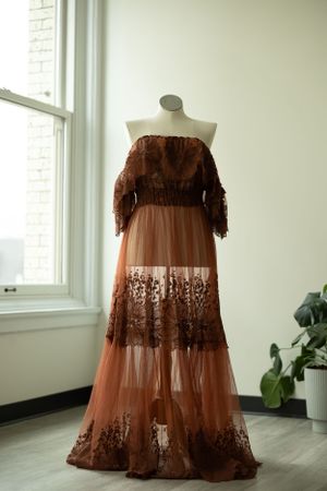 A brown lace off-shoulder formal gown displayed on a mannequin near a window with soft natural lighting.