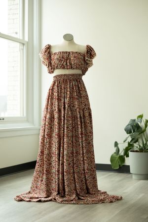 A floral print maternity dress with puff sleeves and empire waist displayed on a mannequin near a window and plant.