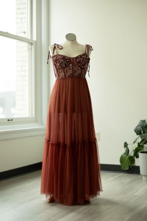 Romantic burgundy tulle evening dress with beaded bodice and tiered skirt displayed on dress form beside window.