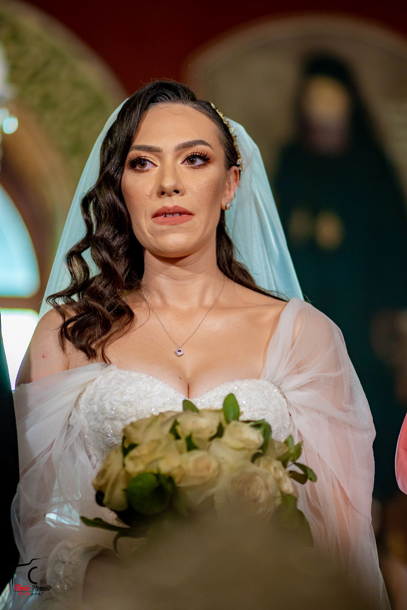 Elegant bride in white veil and lace gown holds cream rose bouquet during wedding ceremony.
