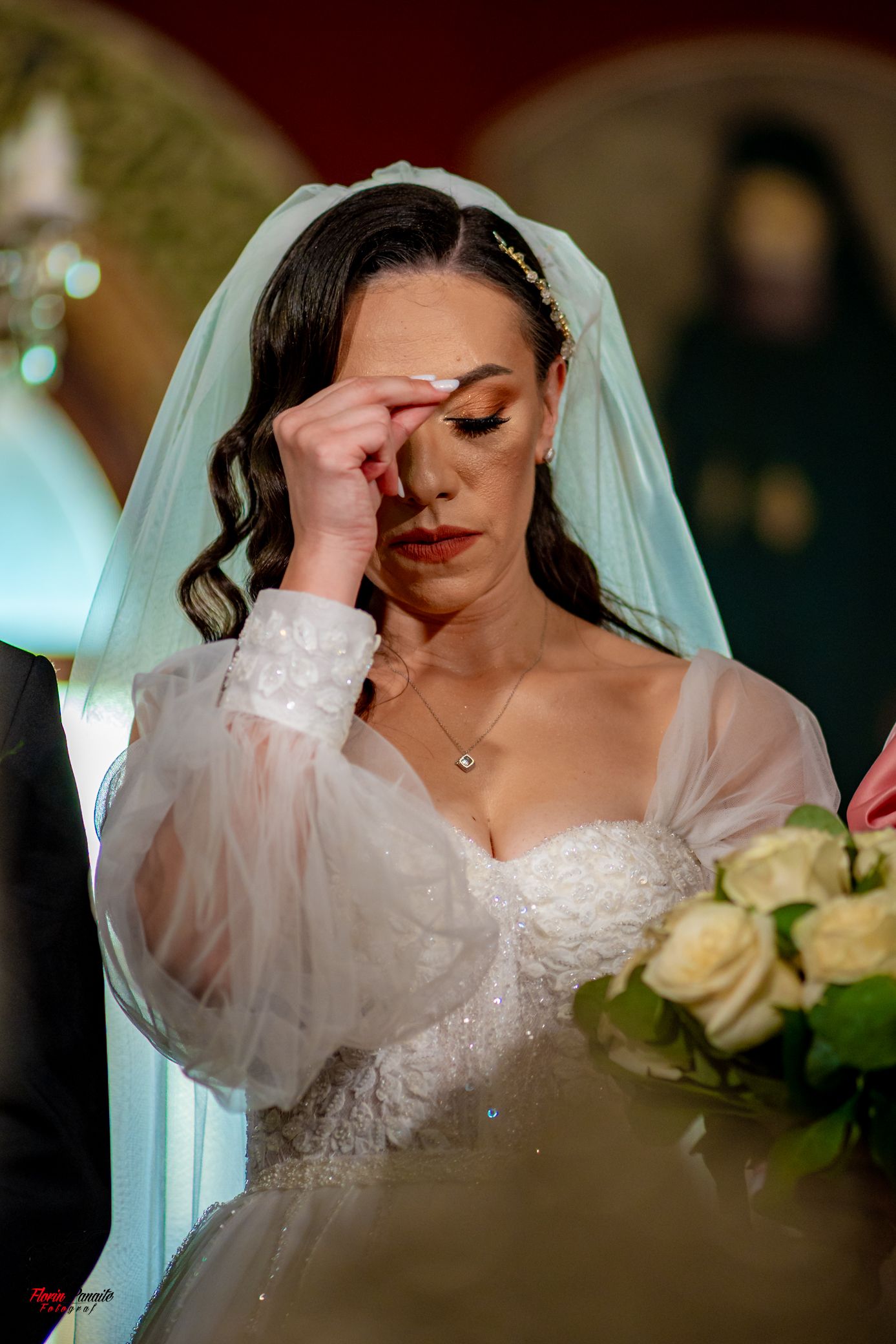 Bride in white lace dress wipes away tears during emotional wedding ceremony moment while holding cream rose bouquet.