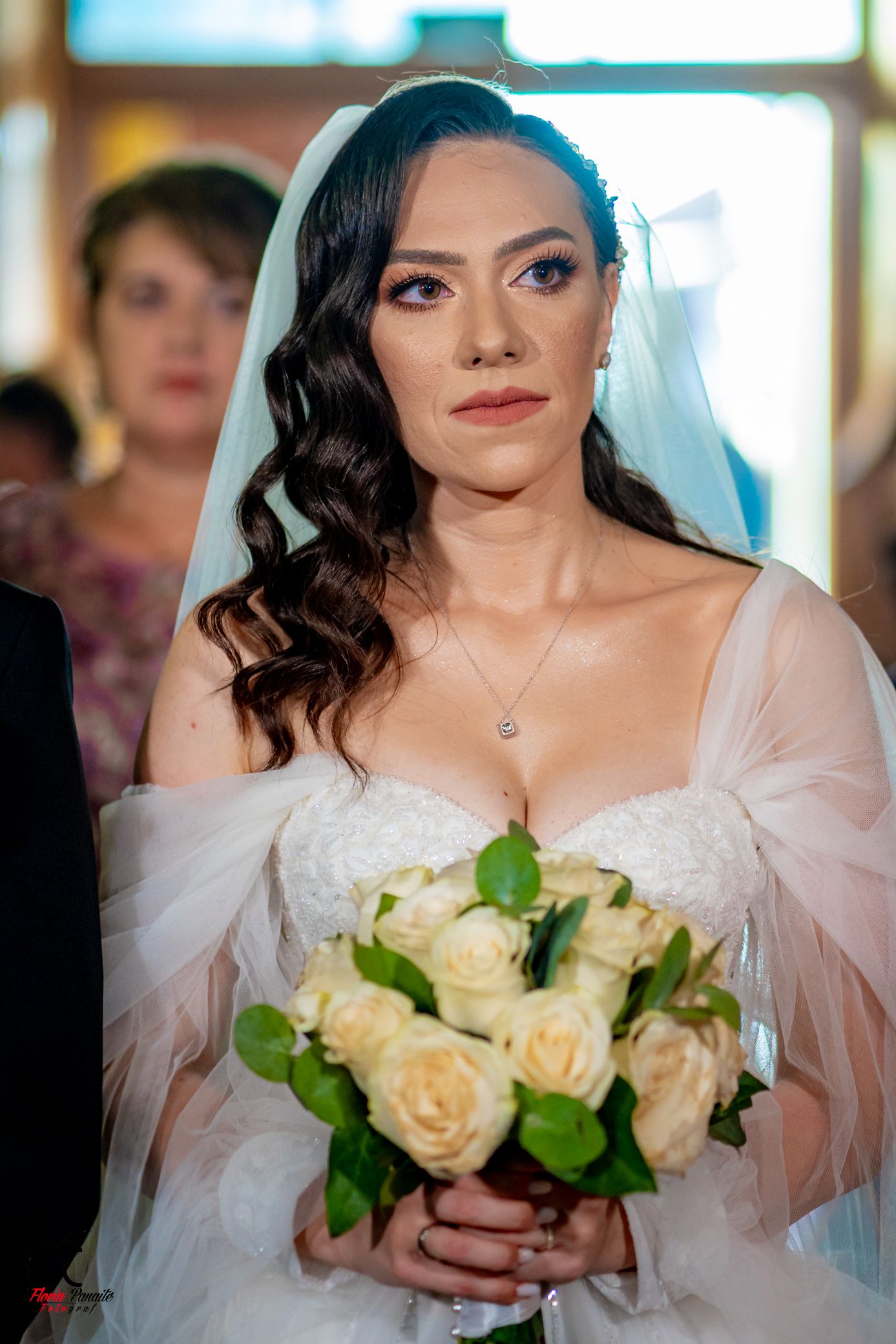 Bride in off-shoulder wedding dress holds cream rose bouquet with greenery during ceremony.