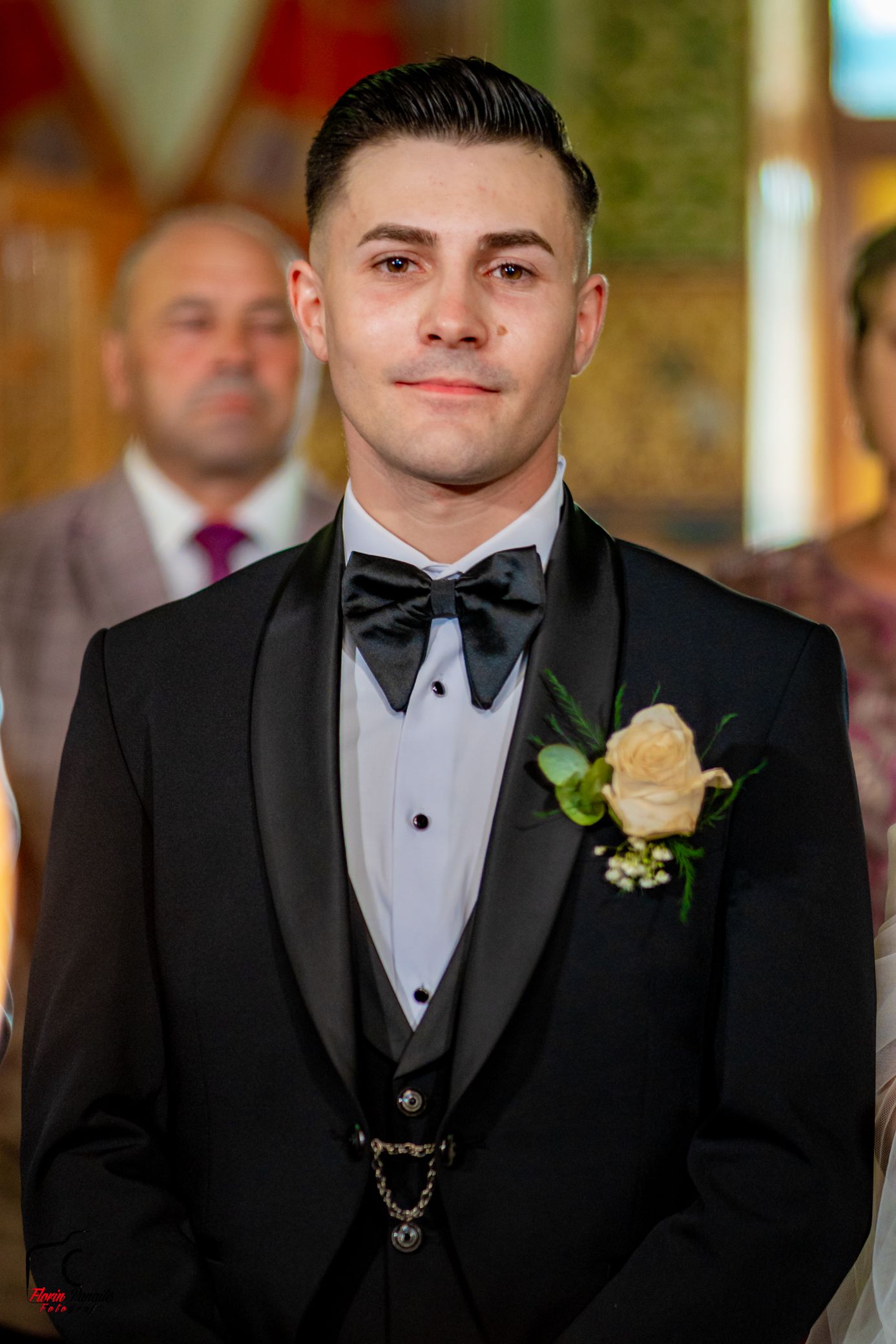 Groom in classic black tuxedo with cream boutonniere stands at wedding altar.